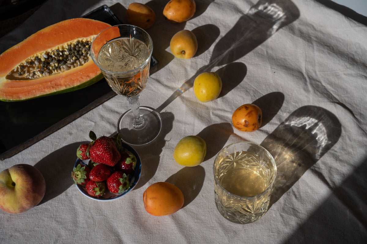 Two crystal glasses filled with cold brew iced tea, surrounded by fresh papaya, strawberries, and small yellow stone fruits on a white tablecloth in bright sunlight.