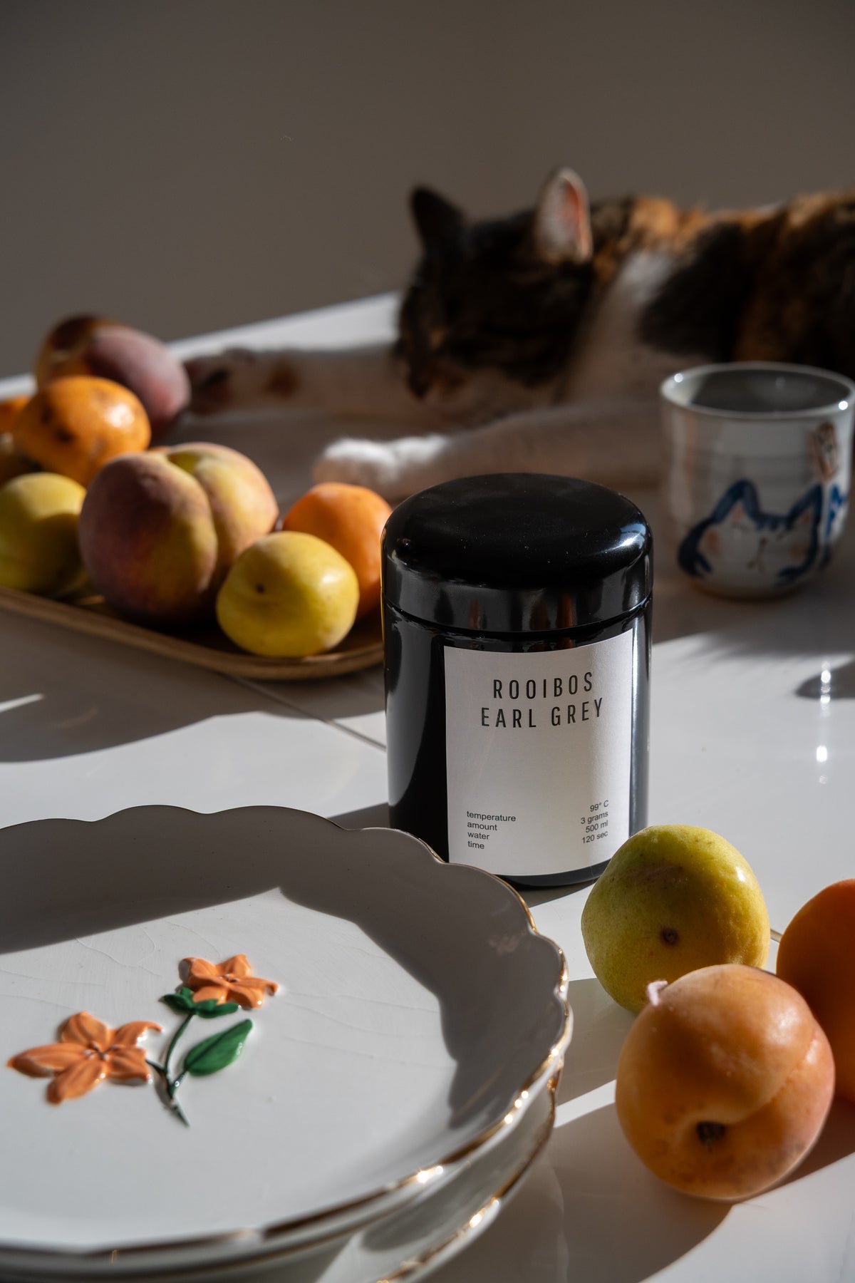 A black tea jar labeled 'ROOIBOS EARL GREY' sits on a white table surrounded by apricots, a ceramic cat mug, and a cat lying in the background on a white table.
