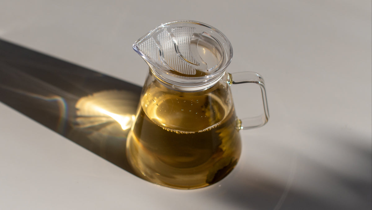 A clear glass pitcher filled with golden yellow liquid, set on a light surface with strong sunlight and a sharp shadow.