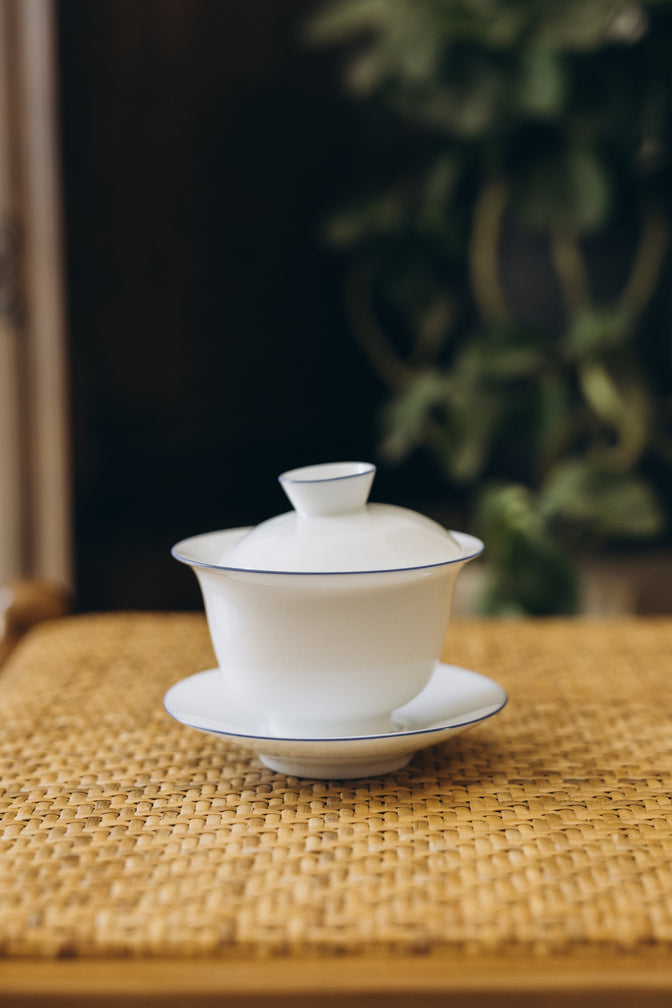 The Cantina white gaiwan with a blue rim on a woven chair.