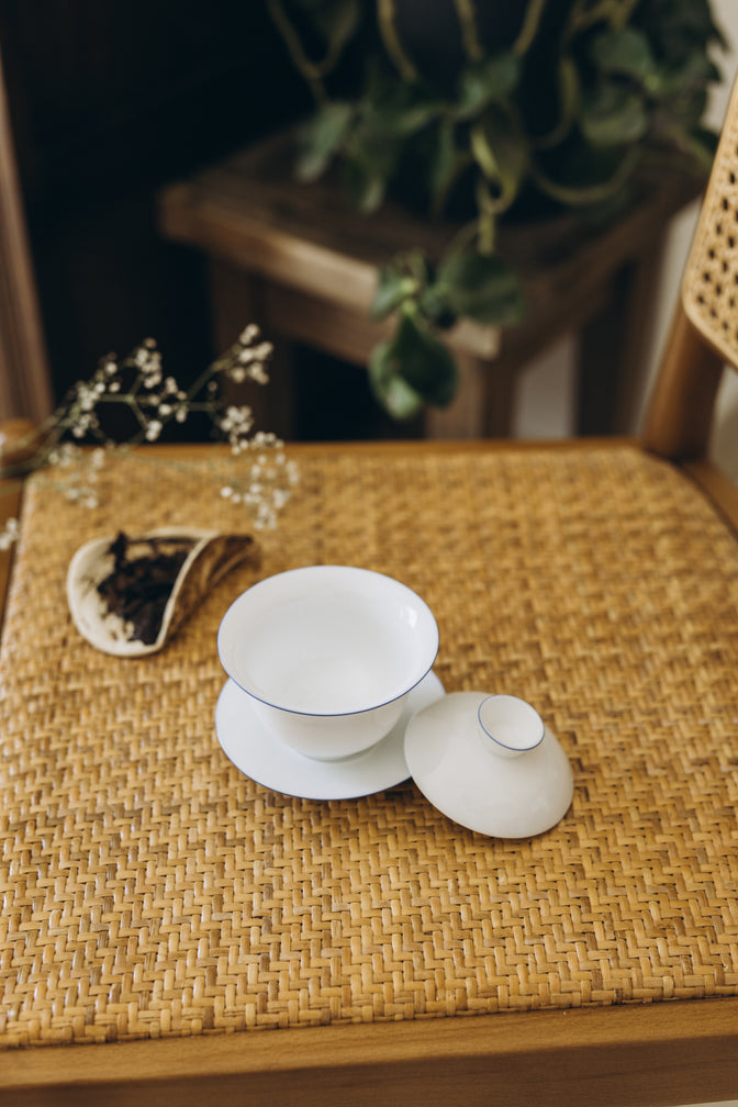 Open Cantina gaiwan with its lid beside the saucer on a woven chair.
