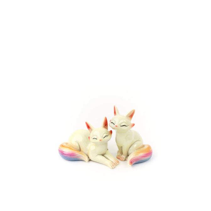 Two cream-colored fox tea pets with rainbow-colored tails.
